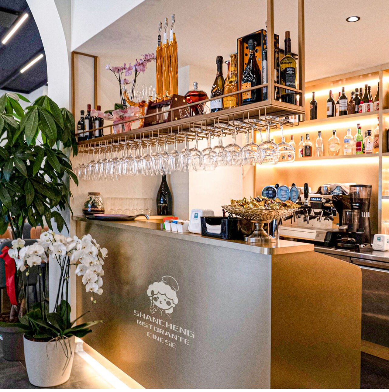 Il bancone bar con bottiglie, calici appesi e un'orchidea bianca.