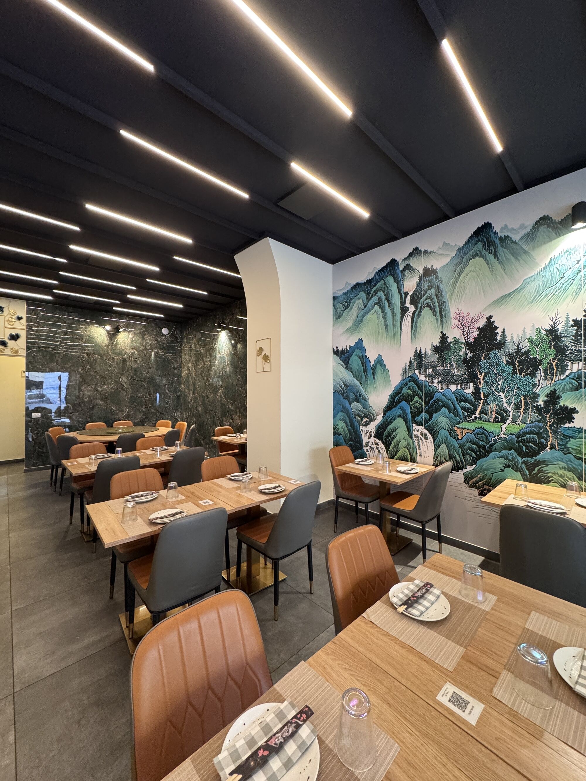 Sala del ristorante: parete interamente dipinta con un paesaggio di montagne cinesi in verde giada, tavoli in legno chiaro e sedie in pelle cuoio.